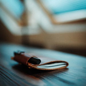 May include: A small, tan leather notebook cover holding a stack of white paper pages, with a black pen inserted into a side loop. The leather has a smooth, natural finish and is folded over the paper, creating a compact, portable writing accessory.
