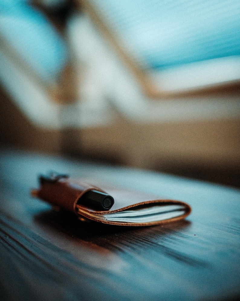 May include: A small, tan leather notebook cover holding a stack of white paper pages, with a black pen inserted into a side loop. The leather has a smooth, natural finish and is folded over the paper, creating a compact, portable writing accessory.