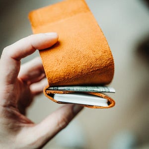 May include: An orange leather wallet, open to reveal a stack of white cards and a folded bill. The wallet has a textured surface and is held in a person's hand. The wallet is a warm, earthy tone.