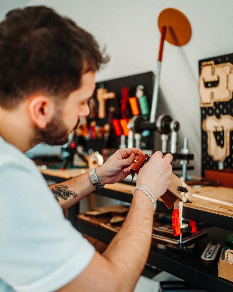 May include: A brown leather piece held in a wooden stitching pony clamp, being hand-stitched with a needle and thread. The leather is secured in the clamp, which is mounted on a wooden workbench, with various leatherworking tools visible nearby.