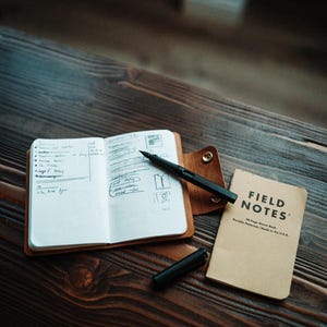 May include: An open notebook with handwritten notes inside a brown leather cover, a black fountain pen with its cap removed, and a tan Field Notes 48-page memo book. The notebook cover features a gold-coloured metal snap button closure.