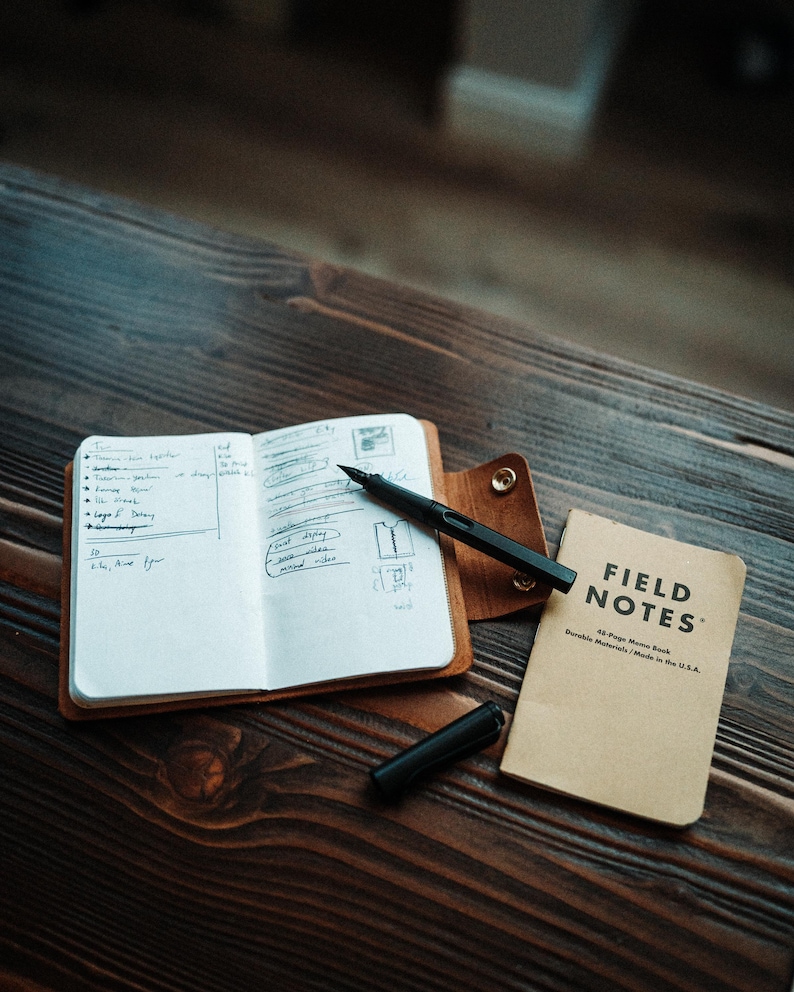 May include: An open notebook with handwritten notes inside a brown leather cover, a black fountain pen with its cap removed, and a tan Field Notes 48-page memo book. The notebook cover features a gold-coloured metal snap button closure.