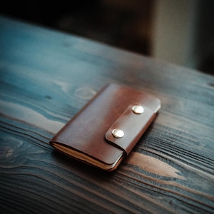 Può includere: Un taccuino in pelle marrone con chiusura a cinturino e due bottoni a pressione color oro. Il taccuino è chiuso e appoggiato su una superficie di legno scuro. La pelle ha una consistenza liscia.