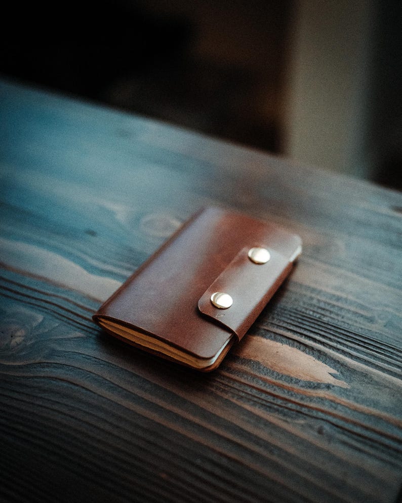 May include: A brown leather notebook cover featuring a wrap-around flap secured by two polished gold-tone metal snap buttons. The rectangular accessory holds a stack of paper pages visible along the side edge, showcasing a smooth finish.