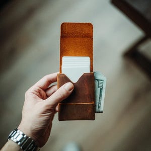 May include: A brown leather wallet with a card slot and a cash pocket. The wallet is open, revealing white cards and a US $100 bill. The wallet is held in a hand, with a silver watch visible on the wrist.