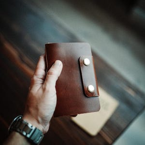 May include: A dark brown leather wallet featuring a vertical flap closure secured by two polished silver-tone metal snap buttons. The rectangular wallet has a smooth, matte finish and rounded corners, showcasing a simple and functional design.