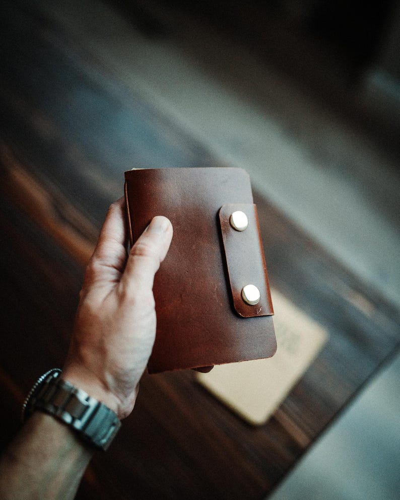 May include: A dark brown leather wallet featuring a vertical flap closure secured by two polished silver-tone metal snap buttons. The rectangular wallet has a smooth, matte finish and rounded corners, showcasing a simple and functional design.