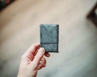 Peut inclure: Un porte-cartes rectangulaire avec une finition gris foncé texturée. L'accessoire présente un design vertical simple avec une fermeture à rabat, mettant en valeur un motif de surface moucheté qui semble uniforme sur le panneau avant.