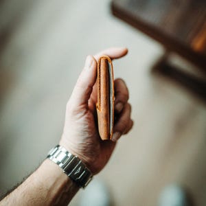 May include: A slim, brown leather wallet held in a hand. The wallet is rectangular and appears to be empty. A silver wristwatch is visible on the wrist. The background is blurred, with a wooden surface and a light-colored floor.