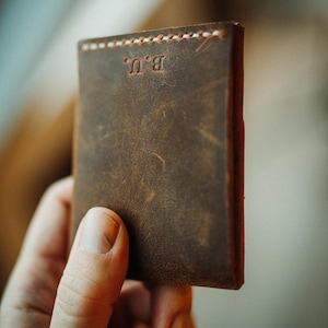 May include: A brown leather wallet with white stitching and the initials "B.U." embossed on the front. The wallet is held in a hand, showcasing its textured surface and compact design. The leather has a distressed appearance.
