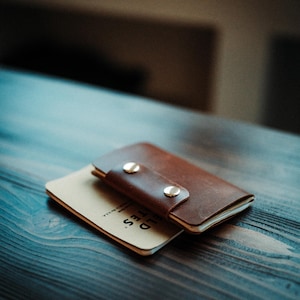 May include: A brown leather notebook cover with two silver-coloured metal snap buttons securing the flap. The cover is wrapped around a tan-coloured notebook with the text FIELD NOTES printed in black on the visible bottom corner of the cover.