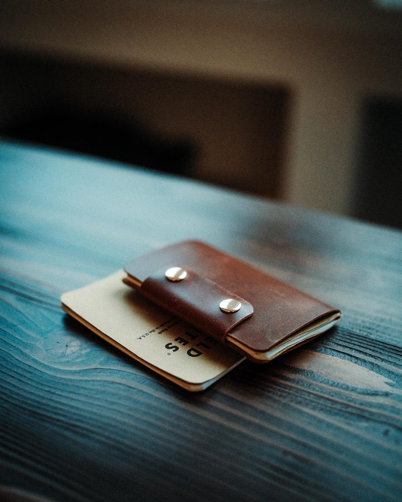 May include: A brown leather notebook cover with two silver-coloured metal snap buttons securing the flap. The cover is wrapped around a tan-coloured notebook with the text FIELD NOTES printed in black on the visible bottom corner of the cover.