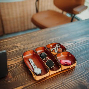 May include: A tan leather valet tray with three distinct compartments secured by metal snaps. The tray holds a silver-tone wristwatch, a pair of black-rimmed sunglasses, a metal keychain, and a small red plastic case for personal items.