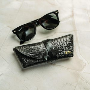 Peut inclure: Lunettes de soleil noires à verres foncés placées au-dessus d'un étui à lunettes en cuir noir à texture crocodile. L'étui est doté d'une fermeture à lanière en cuir et des initiales B.U. gaufrées en doré dans le coin inférieur droit.