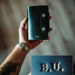 May include: A black leather wallet featuring a vertical flap secured by two silver-toned metal snaps. The front surface is embossed with the letters B.U. in a serif font. The wallet has a smooth texture and a rectangular shape with rounded corners.