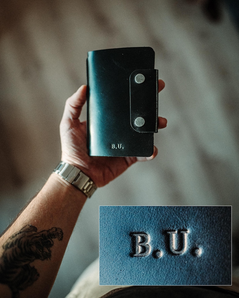 May include: A black leather wallet featuring a vertical flap secured by two silver-toned metal snaps. The front surface is embossed with the letters B.U. in a serif font. The wallet has a smooth texture and a rectangular shape with rounded corners.