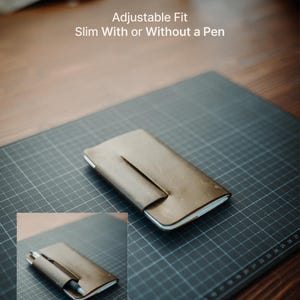 May include: A brown leather notebook cover with a wrap-around closure flap. The text "Adjustable Fit Slim With or Without a Pen" is visible. An inset shows the cover holding a pen, demonstrating its function as a slim, adjustable notebook holder.