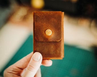 May include: A rectangular brown leather card holder featuring a smooth, matte finish and a single centered brass-colored metal snap button closure. The leather has visible natural grain textures and stitched edges, forming a compact, vertical pocket.