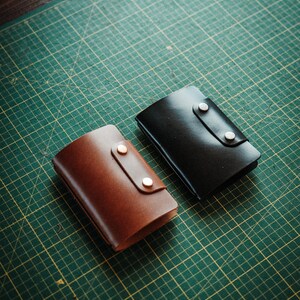 May include: Two rectangular leather wallets, one in a rich brown and the other in a deep black, each featuring a smooth finish and a secure flap closure held in place by two metallic snap buttons positioned vertically on the front panel.