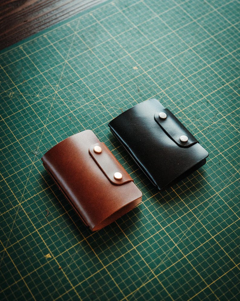 May include: Two rectangular leather wallets, one in a rich brown and the other in a deep black, each featuring a smooth finish and a secure flap closure held in place by two metallic snap buttons positioned vertically on the front panel.