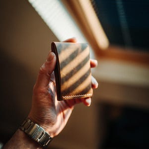 May include: A brown leather wallet with visible stitching, held in a hand. The wallet is rectangular and appears to be made of high-quality leather. The image is lit by sunlight, creating shadows across the wallet.