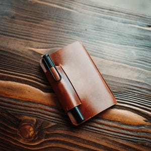 May include: A rectangular brown leather notebook cover featuring a built-in pen loop on the front flap. A matte black fountain pen with a wire clip is inserted into the loop, resting securely against the smooth, polished surface of the leather.