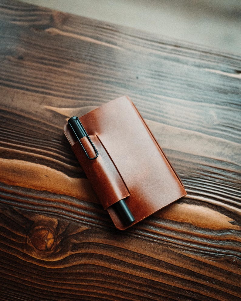 May include: A rectangular brown leather notebook cover featuring a built-in pen loop on the front flap. A matte black fountain pen with a wire clip is inserted into the loop, resting securely against the smooth, polished surface of the leather.