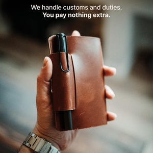May include: A brown leather notebook cover featuring an integrated pen loop holding a black fountain pen. The text We handle customs and duties. You pay nothing extra. is visible at the top of the image. The cover has a smooth, matte finish.