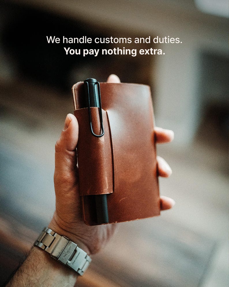May include: A brown leather notebook cover featuring an integrated pen loop holding a black fountain pen. The text We handle customs and duties. You pay nothing extra. is visible at the top of the image. The cover has a smooth, matte finish.