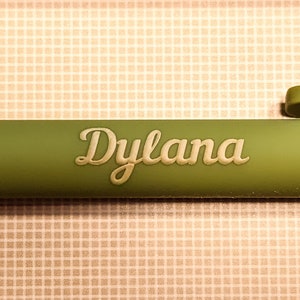 May include: Close-up of a green pen with the name "Dylana" in a cream-colored, cursive font. The pen is set against a patterned background of small squares in shades of beige and cream.