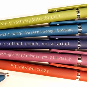 May include: A collection of colorful pens with humorous phrases in white text. The pens are in shades of green, blue, purple, pink, and orange. The phrases include "Ain't nothing Soft about it" and "Pitches be crazy."