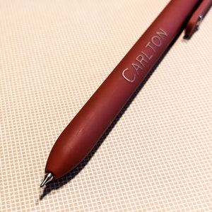May include: A burgundy pen with the name "CARLTON" engraved on the barrel. The pen has a silver clip and a fine point. The pen is lying on a beige, textured surface.