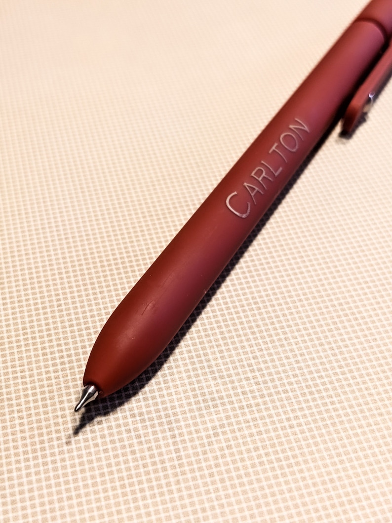 May include: A burgundy pen with the name "CARLTON" engraved on the barrel. The pen has a silver clip and a fine point. The pen is lying on a beige, textured surface.