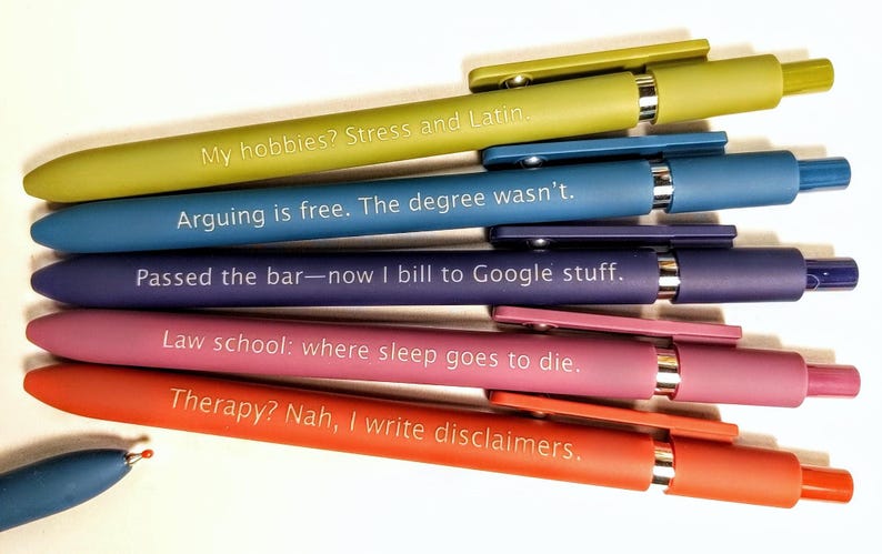 May include: A collection of colorful pens with humorous phrases printed on them. The pens are in shades of green, blue, purple, pink, and orange. The text includes phrases like "My hobbies? Stress and Latin" and "Therapy? Nah, I write disclaimers."