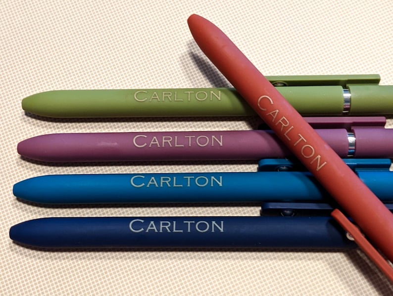 May include: A collection of colorful pens in shades of green, purple, blue, and red. Each pen has the name "CARLTON" printed on it. The pens are arranged on a light beige surface.