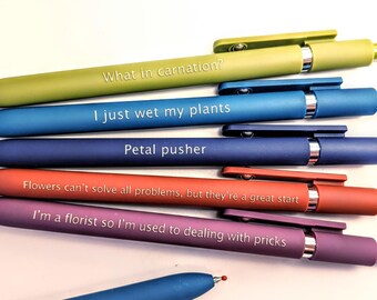 May include: A selection of pens in various colours, each with a different pun printed on them. The pens are in shades of green, blue, red, and purple. Phrases include "What in carnation?" and "I'm a florist so I'm used to dealing with pricks."