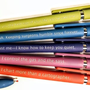 May include: A collection of colorful pens with humorous phrases printed on them. The pens are in shades of green, blue, purple, pink, and red. The text includes phrases like "I give you the best nap of your life" and "I control the gas and the sass."