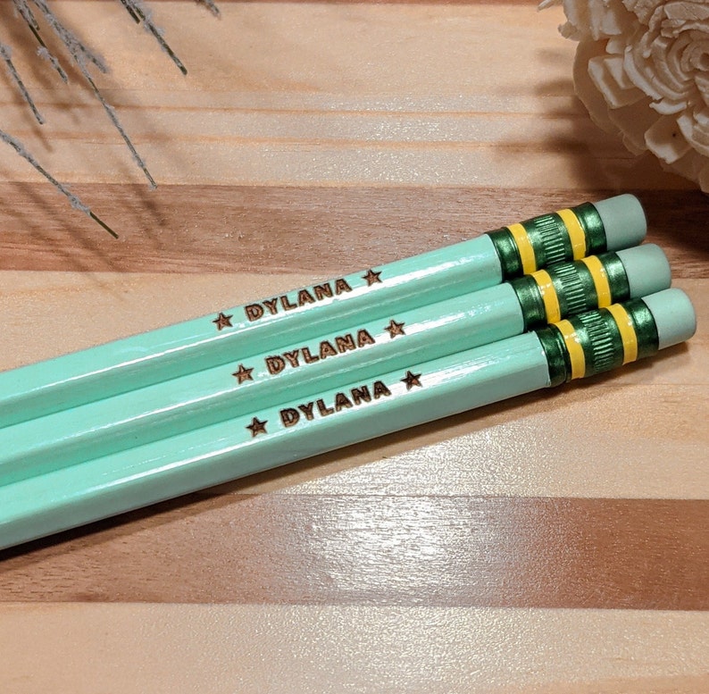 May include: Three mint green pencils, each engraved with the name "DYLANA" and a star. The pencils feature green and yellow detailing near the eraser and are arranged diagonally on a wooden surface.