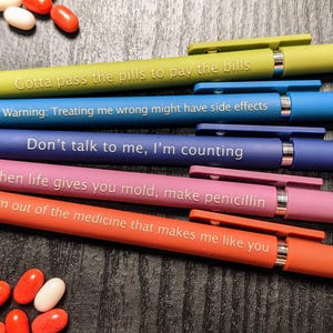 Pode incluir: Cinco canetas coloridas com ditos humorísticos sobre pílulas e medicamentos. Os ditos são: "Gotta pass the pills to pay the bills", "Warning: Treating me wrong might have side effects", "Don't talk to me, I'm counting", "When life gives you mold, make penicillin", e "I am out of the medicine that makes me like you".
