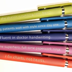 Puede incluir: Un conjunto de bolígrafos coloridos con frases humorísticas impresas. Los bolígrafos son de color verde, azul, morado, rosa y rojo. Cada bolígrafo tiene un clip y una punta plateados. Los textos incluyen frases como "I fix charts, not hearts."