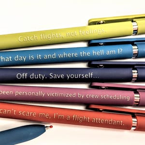 May include: A collection of colorful pens with humorous phrases in white text. The pens are in shades of green, blue, navy, purple, and red. The phrases include "Catch flights, not feelings" and "You can't scare me, I'm a flight attendant."
