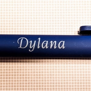 May include: A navy blue pen with the name "Dylana" engraved in a delicate, cursive font. The pen has a clip and a smooth, matte finish. The background is a light tan, checkered pattern.