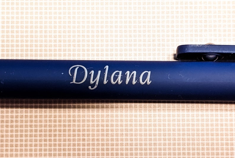 May include: A navy blue pen with the name "Dylana" engraved in a delicate, cursive font. The pen has a clip and a smooth, matte finish. The background is a light tan, checkered pattern.