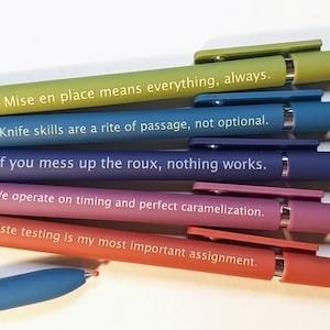 Könnte beinhalten: Ein Set aus sechs einziehbaren Kugelschreibern in verschiedenen Farben wie Limettengrün, Blau, Lila, Pink und Orange. Jeder Stift trägt ein einzigartiges kulinarisches Zitat in weißer Schrift auf dem Gehäuse, wie "Mise en place means everything, always" und "Taste testing is my most important assignment."