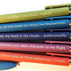 May include: An assortment of colorful pens with humorous phrases printed on them. The pens are in shades of green, blue, purple, pink, and orange. The text includes phrases like "Coffee, checklists, repeat" and "Turbulence just adds character to the flight."
