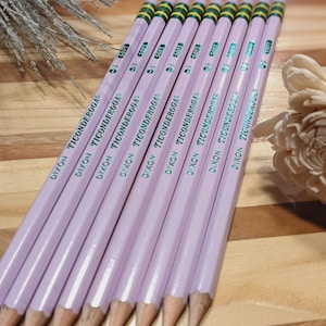 May include: A group of light purple Dixon Ticonderoga pencils. Each pencil features a green and yellow striped eraser end and the text "DIXON TICONDEROGA". The pencils are displayed on a wooden surface.