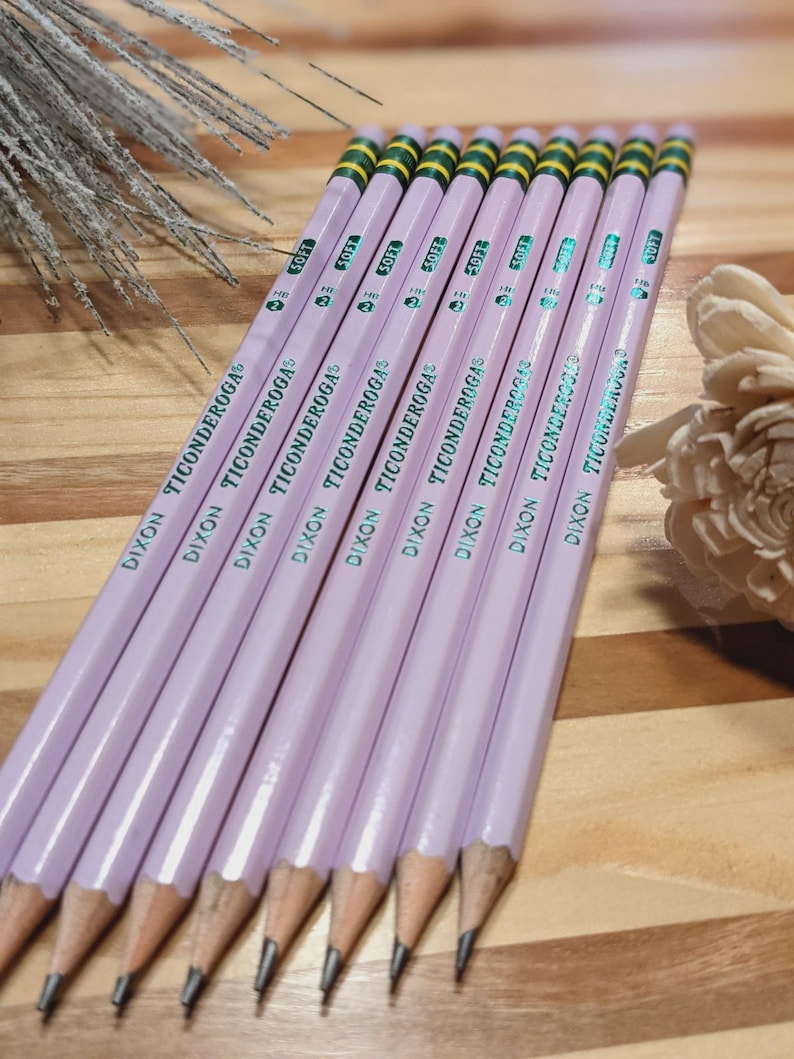 May include: A group of light purple Dixon Ticonderoga pencils. Each pencil features a green and yellow striped eraser end and the text "DIXON TICONDEROGA". The pencils are displayed on a wooden surface.