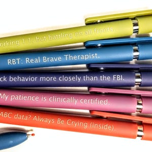 May include: A selection of colourful pens with witty phrases printed on them. The pens are in shades of green, blue, purple, pink, and red. The text includes phrases like "Working 1:1, but battling on all fronts" and "My patience is clinically certified."