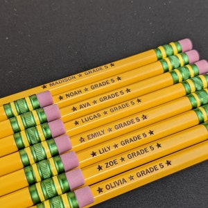 May include: A selection of yellow pencils with pink erasers and green bands. Each pencil is inscribed with a name and the text "GRADE 5" in black. The names include Madison, Noah, Ava, Lucas, Emily, Lily, Zoe, and Olivia.