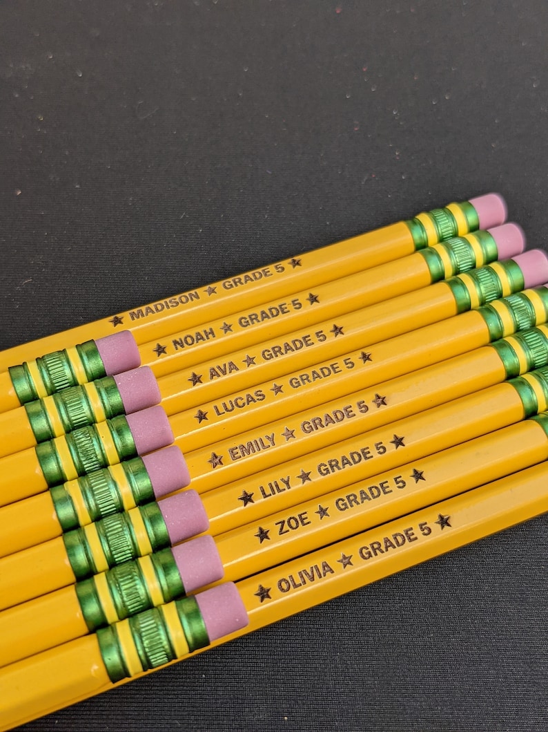 May include: A selection of yellow pencils with pink erasers and green bands. Each pencil is inscribed with a name and the text "GRADE 5" in black. The names include Madison, Noah, Ava, Lucas, Emily, Lily, Zoe, and Olivia.